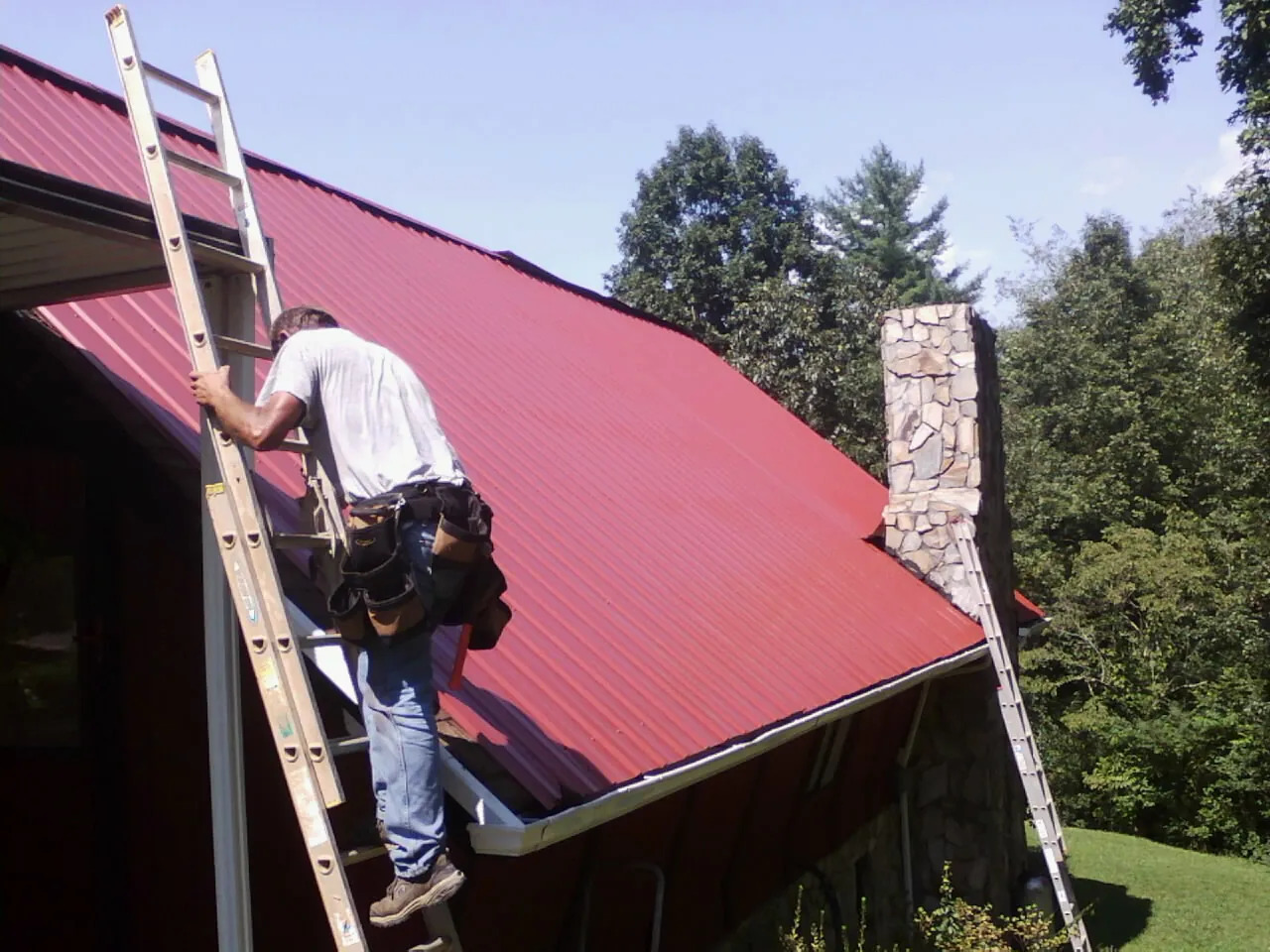 Roofer on a ladder beside a red metal roof and stone chimney — Soffit & Fascia Repair by Roofing Brothers in Bowerston